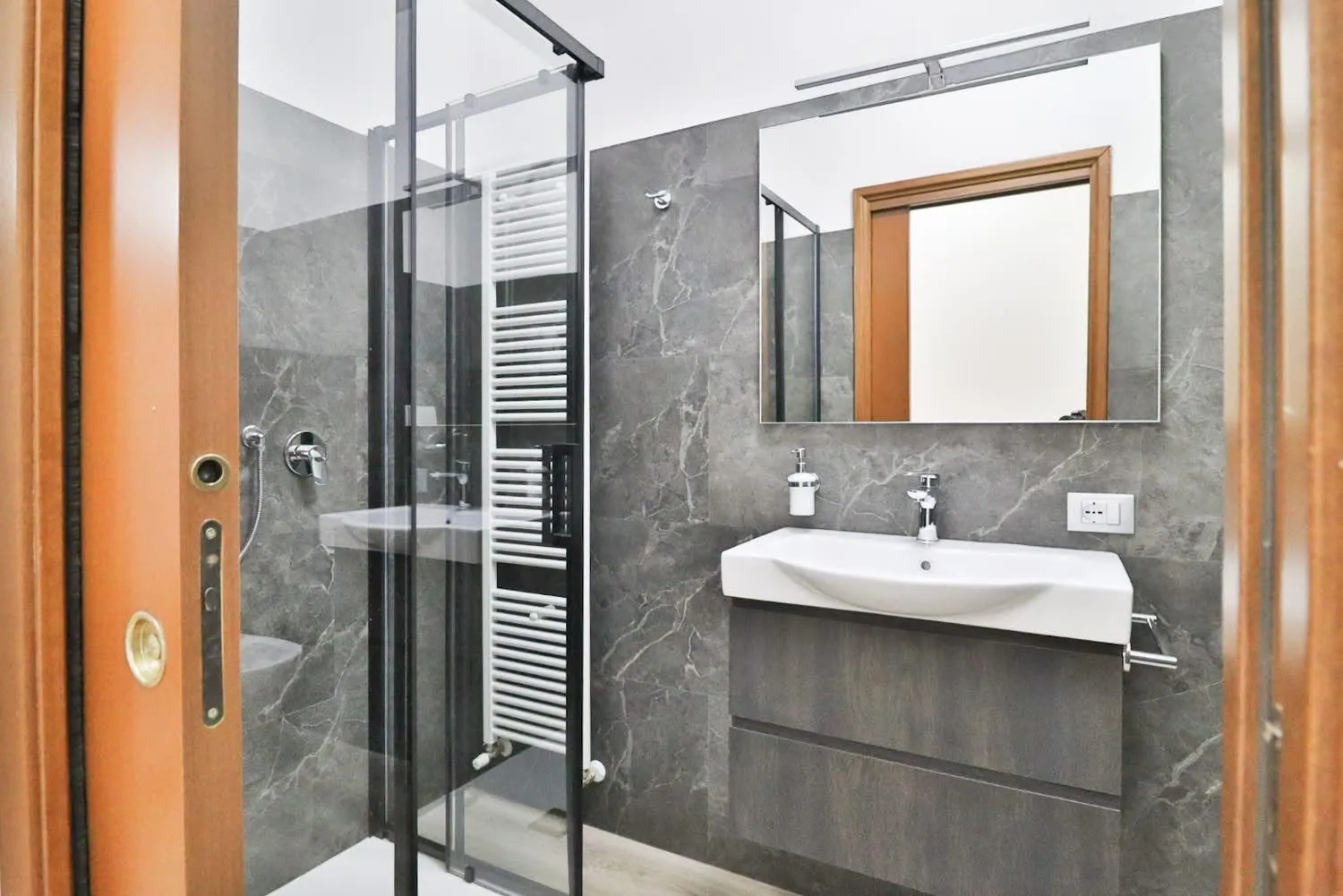 Bagno con doccia in vetro, lavandino e rivestimento in marmo grigio